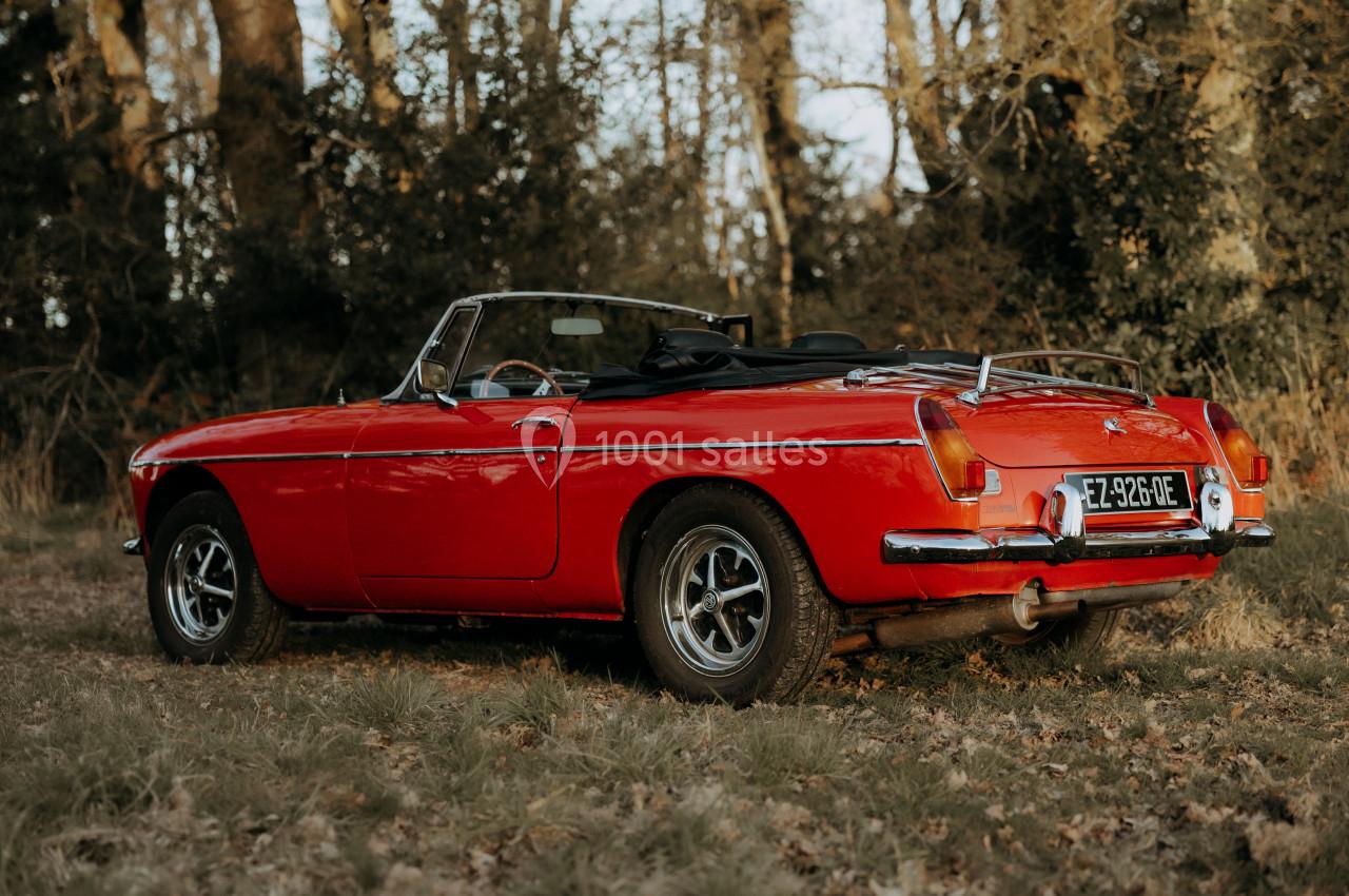 Voiture ancienne décapotable rouge garée sur une pelouse, entourée d'arbres en arrière-plan.