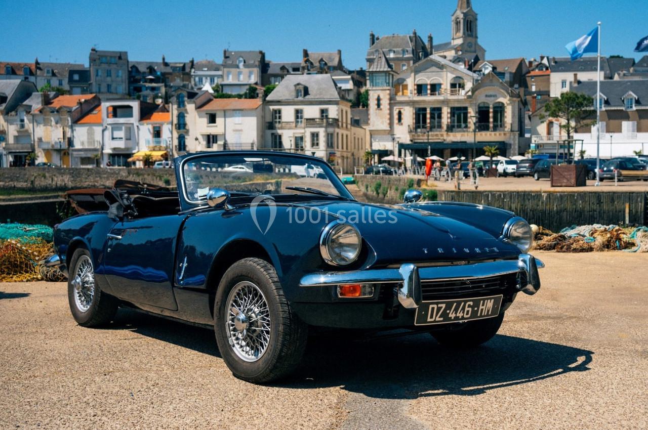 Voiture décapotable classique noire stationnée sur un quai, avec des bâtiments et une église en arrière-plan.
