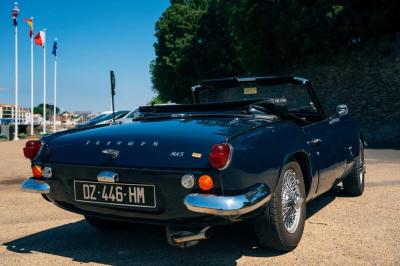 Miniature Voitures Passion #4 Roue avant d'une voiture vintage beige avec un enjoliveur chromé, stationnée sur un sol gravillonné.