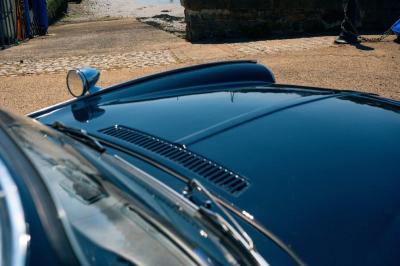 Miniature Voitures Passion #6 Roue avant d'une voiture vintage beige avec un enjoliveur chromé, stationnée sur un sol gravillonné.