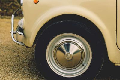 Miniature Voitures Passion #10 Roue avant d'une voiture vintage beige avec un enjoliveur chromé, stationnée sur un sol gravillonné.