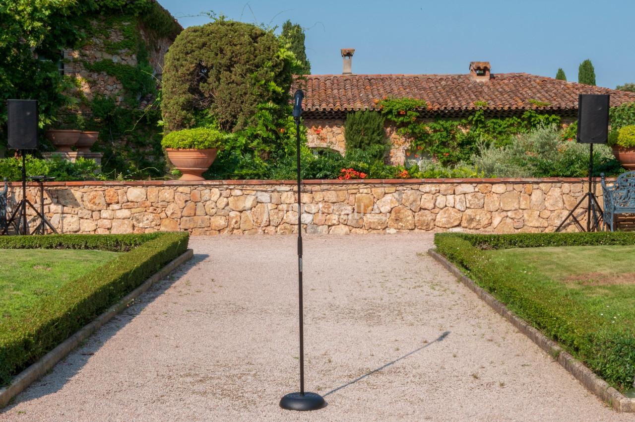Microphone sur pied installé dans un jardin avec allée gravillonnée, haies taillées et mur en pierre en arrière-plan.
