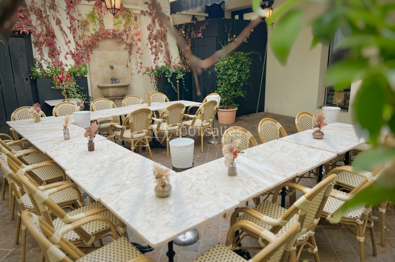 Terrasse extérieure avec tables en marbre disposées en U, chaises en rotin, plantes et fontaine murale en arrière-plan.