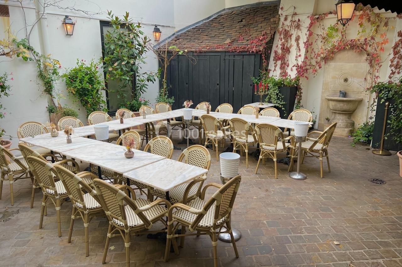 Terrasse pavée avec tables en marbre, chaises en rotin, plantes grimpantes et une fontaine murale en arrière-plan.