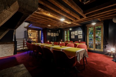 Salle à manger chaleureuse avec poutres apparentes, tables dressées, chaises rouges et cheminée en pierre.
