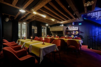 Salle à manger chaleureuse avec poutres apparentes, tables dressées, chaises rouges et cheminée en pierre.