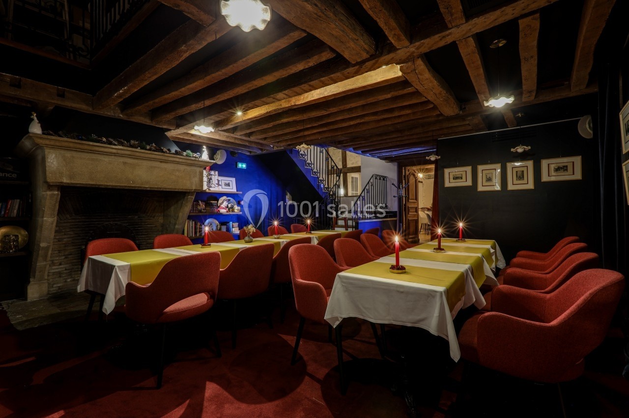 Salle de restaurant chaleureuse avec tables dressées, chaises rouges, poutres apparentes et éclairage tamisé.