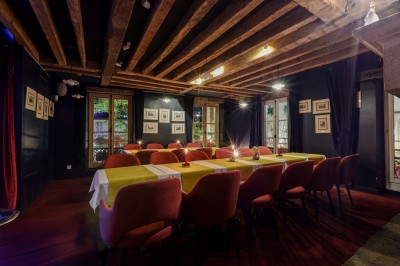 Salle à manger chaleureuse avec poutres apparentes, tables dressées, chaises rouges et cheminée en pierre.