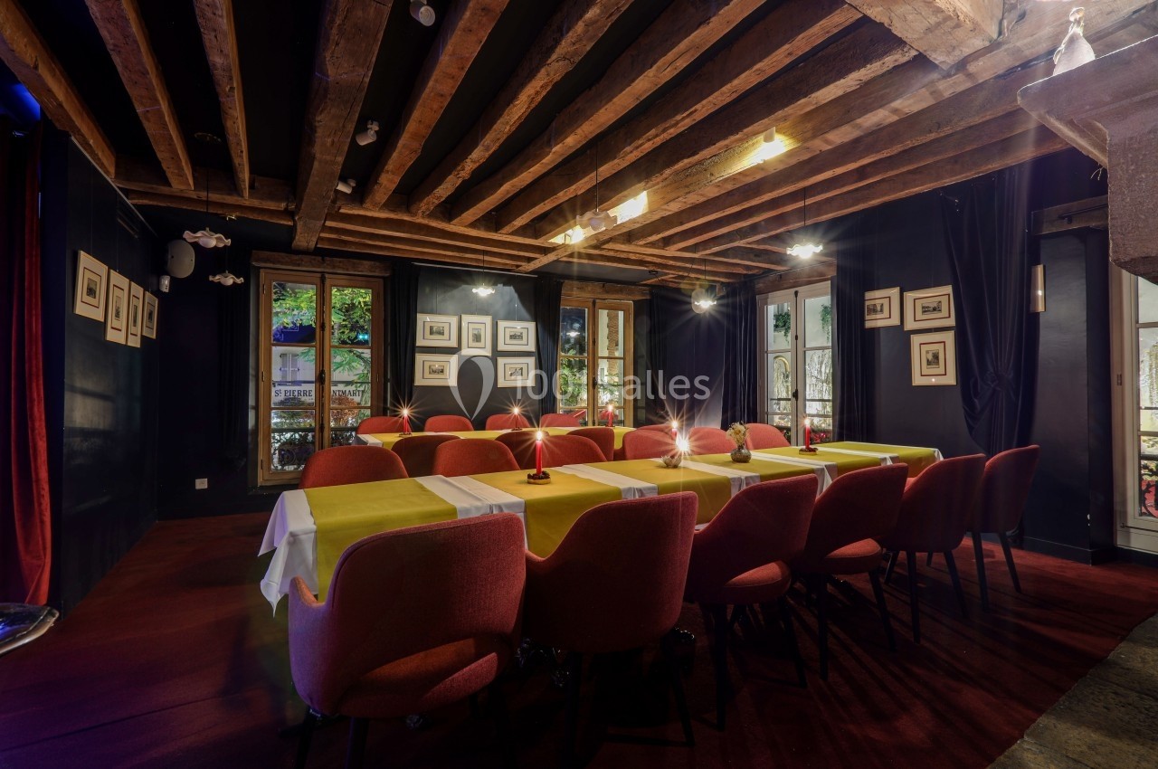 Salle de réunion chaleureuse avec poutres apparentes, tables dressées et chaises rouges, éclairée par des fenêtres.