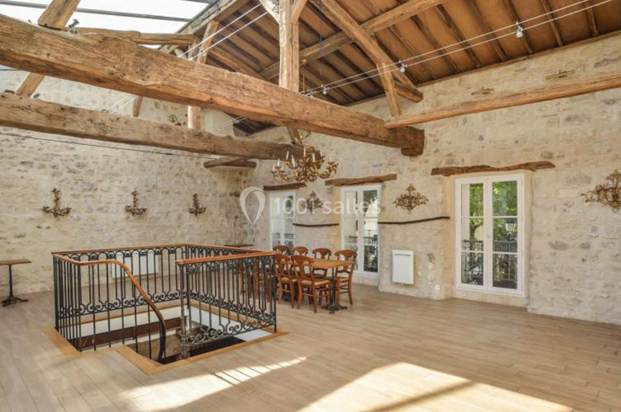 Salle lumineuse avec poutres apparentes, murs en pierre, parquet clair, table en bois et rambarde en fer forgé.