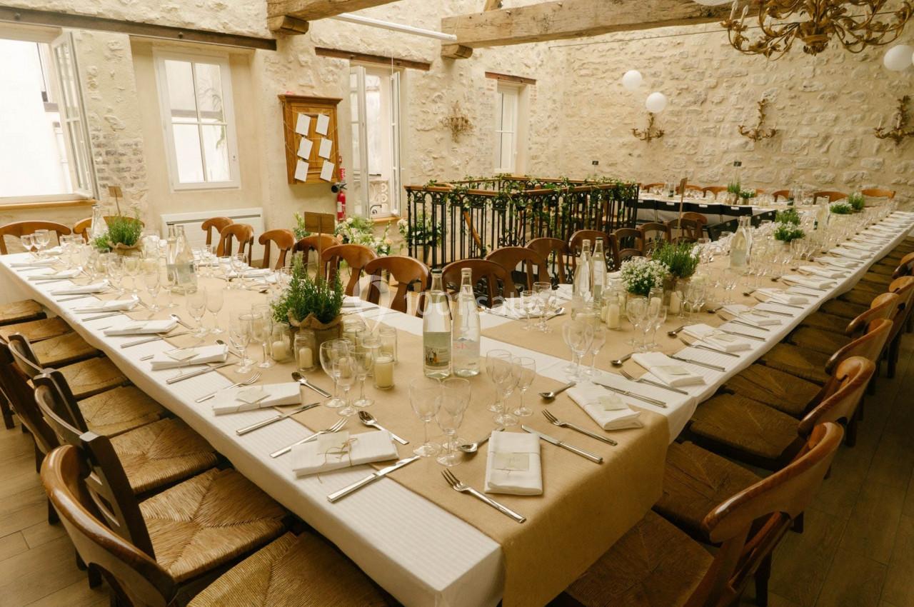 Grande table en U dressée pour un repas, avec nappes beige, vaisselle élégante et décoration végétale dans une salle en…