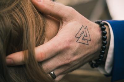 Main avec un tatouage de triangles entrelacés, posée sur des cheveux bruns, bracelet en perles noires visible.