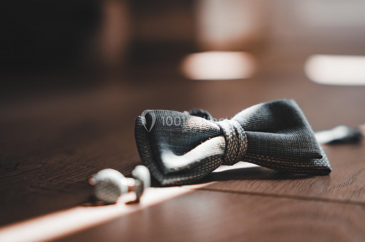 Nœud papillon gris et boutons de manchette posés sur un sol en bois, éclairés par une lumière douce.