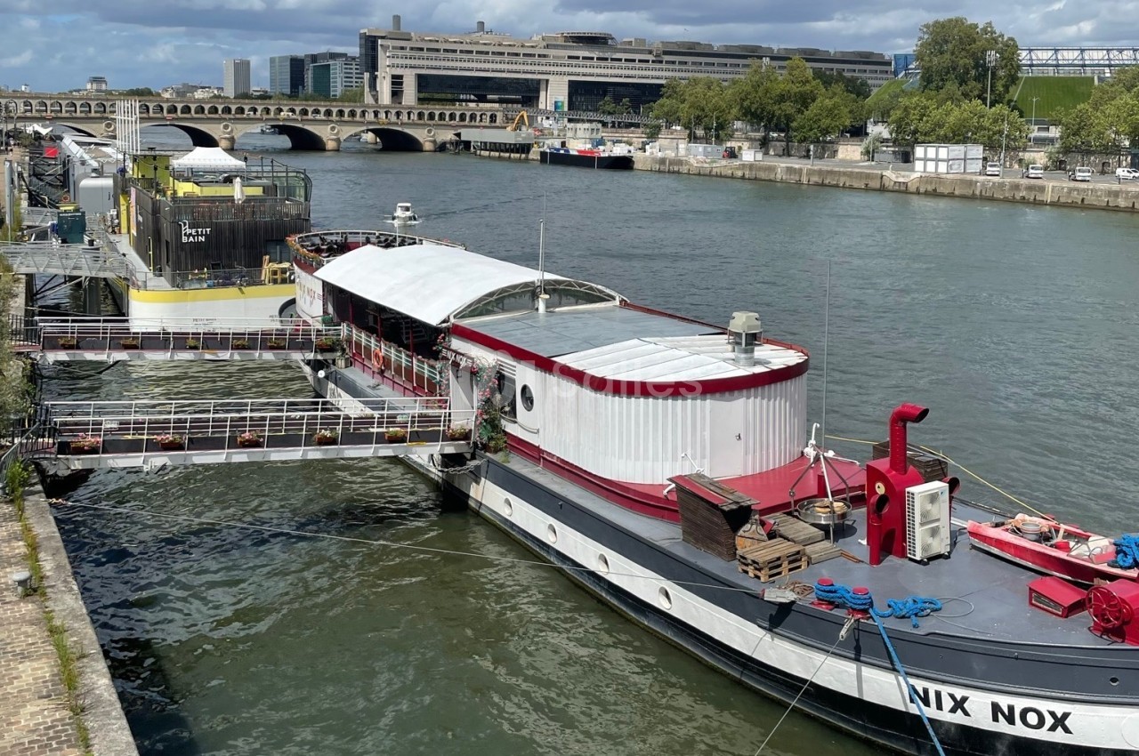 Péniche blanche et rouge amarrée sur la Seine avec terrasse couverte et pont de Paris en arrière-plan.