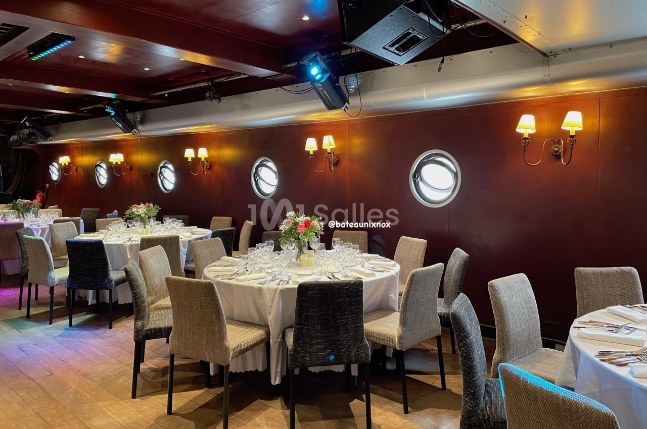 Salle de réception dressée avec tables rondes en nappe blanche, chaises beiges et noires, murs bordeaux, hublots ronds, appliques dorées et éclairage tamisé.
