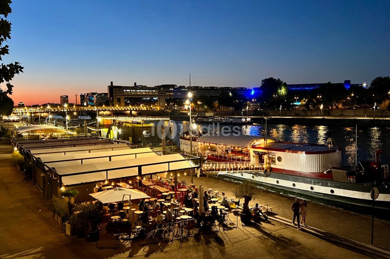 Terrasse au bord de l'eau de nuit avec convives attablés, structure flottante et bâtiments illuminés en arrière-plan.