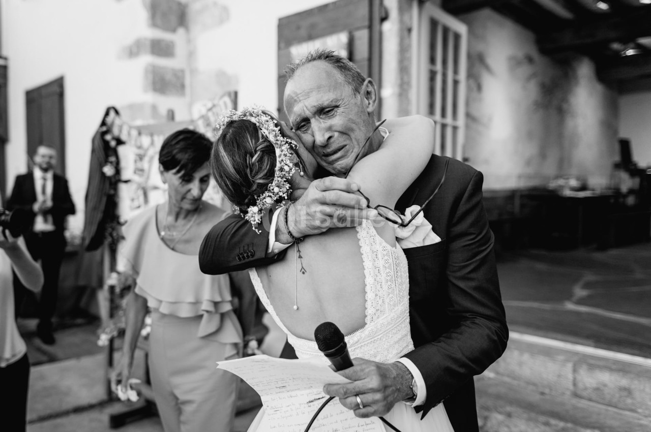 Un homme ému enlace une femme en robe de mariée, tenant un micro, dans un moment de tendresse lors d'une cérémonie.