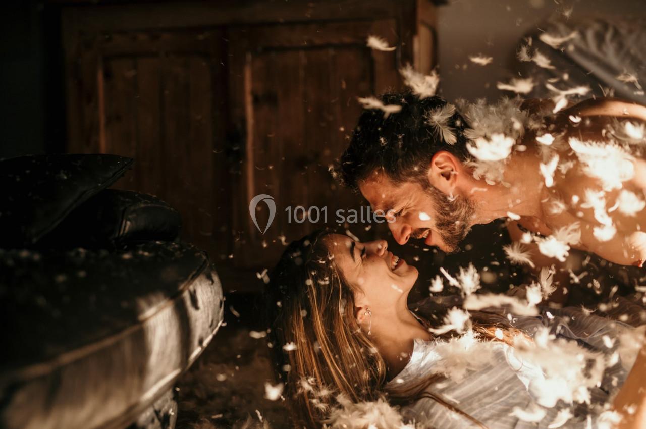 Un couple souriant allongé au sol entouré de plumes dans une ambiance chaleureuse et lumineuse.