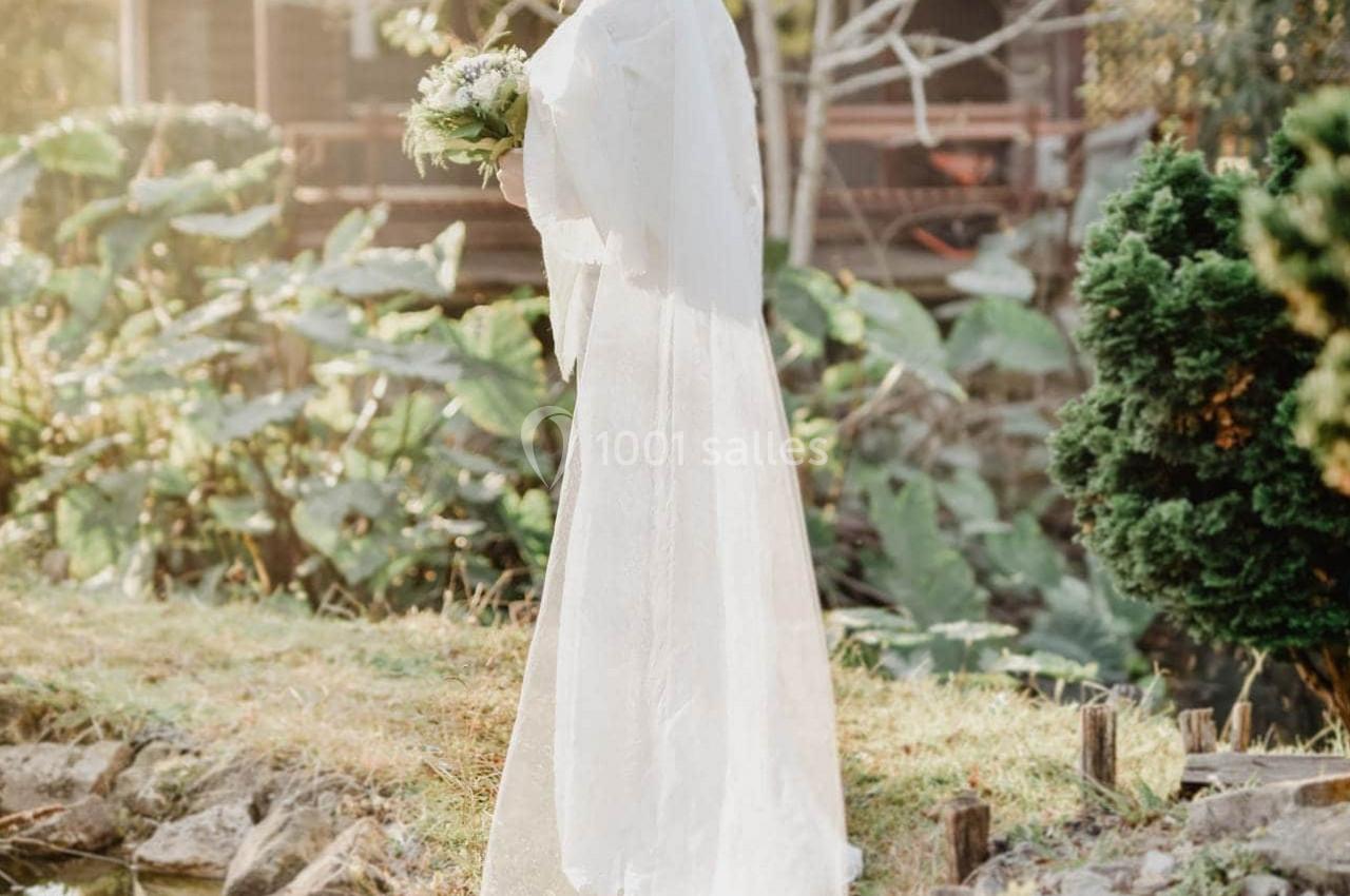 Une femme en robe de mariée blanche tient un bouquet près d'un étang, devant une maison en bois et un jardin verdoyant.