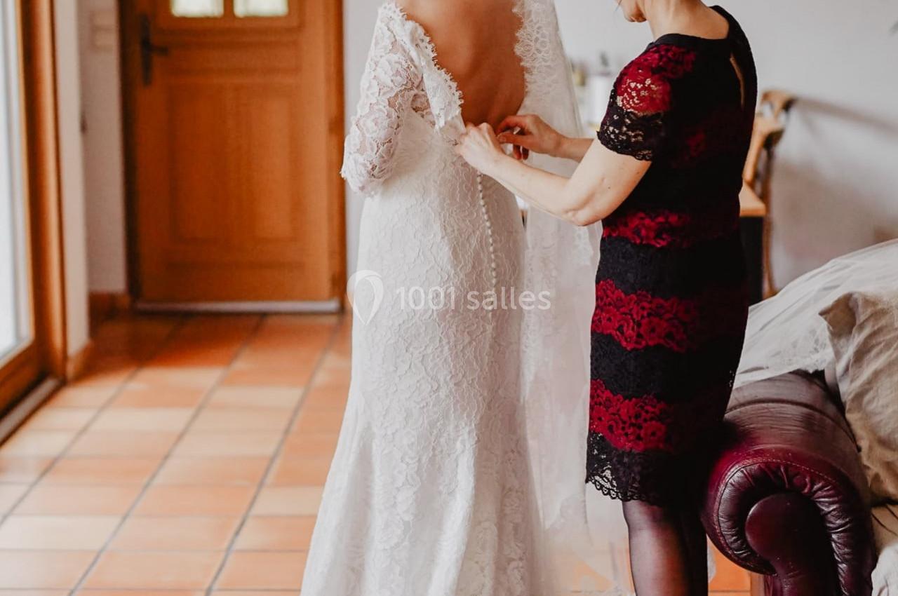 Une femme aide une mariée à ajuster sa robe blanche dans une pièce lumineuse avec un sol en carrelage.