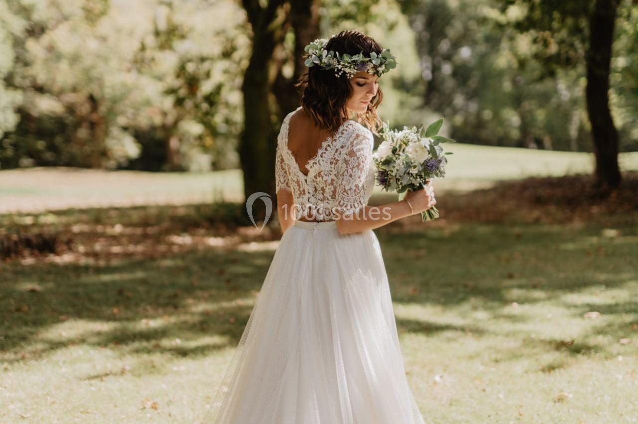 Une femme en robe de mariée blanche tient un bouquet, debout dans un parc ensoleillé entouré d'arbres.