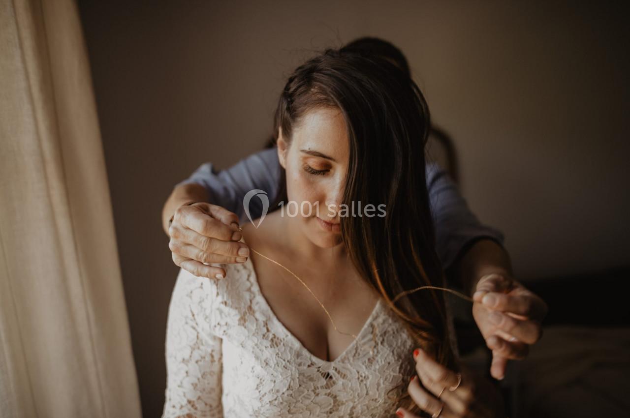 Une personne ajuste un collier autour du cou d'une femme portant une robe en dentelle blanche.