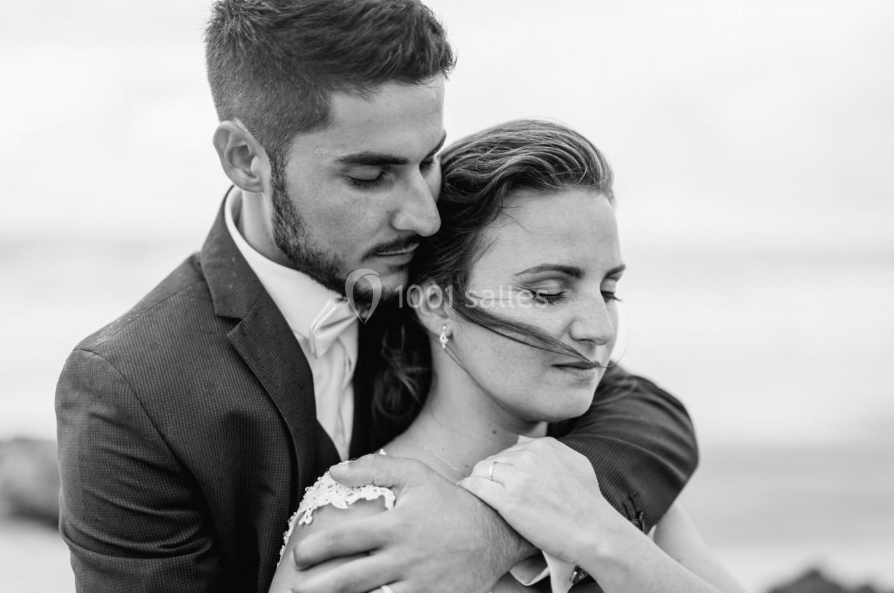 Un couple en tenue de mariage s'enlace tendrement en noir et blanc, avec un arrière-plan flou de plage.