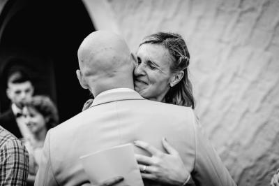 Un couple assis lors d'une cérémonie, l'homme ému essuie une larme tandis que la femme le regarde avec tendresse.
