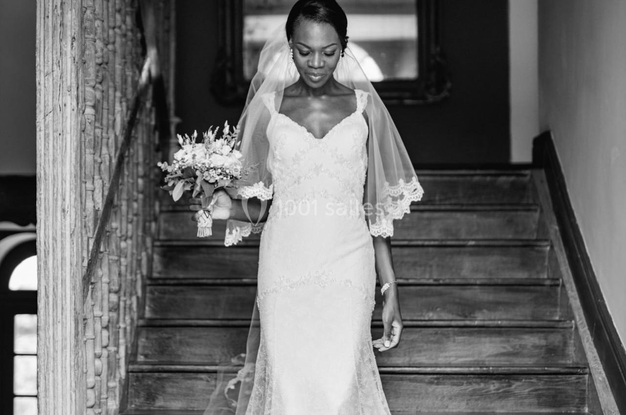 Une mariée en robe blanche et voile descend un escalier en bois, tenant un bouquet de fleurs.