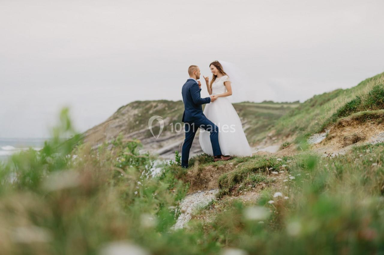 Un couple en tenue de mariage pose sur une colline verdoyante avec un paysage côtier en arrière-plan.