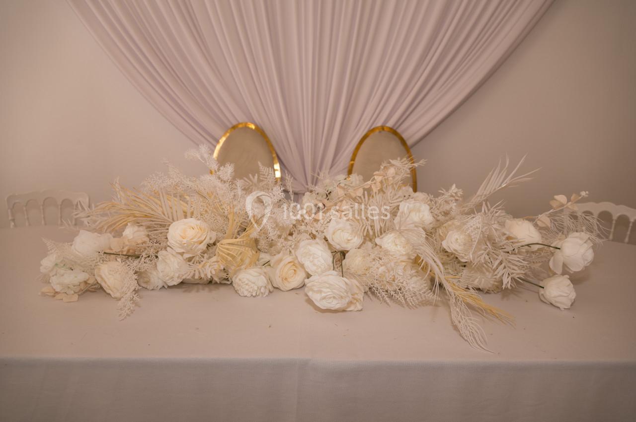 Arrangement floral blanc avec roses et feuillages secs, posé sur une table blanche devant un fond drapé clair.