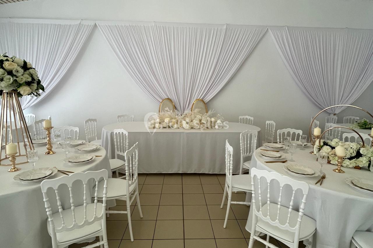 Salle de réception décorée en blanc avec des tables rondes, des chaises élégantes et des arrangements floraux.
