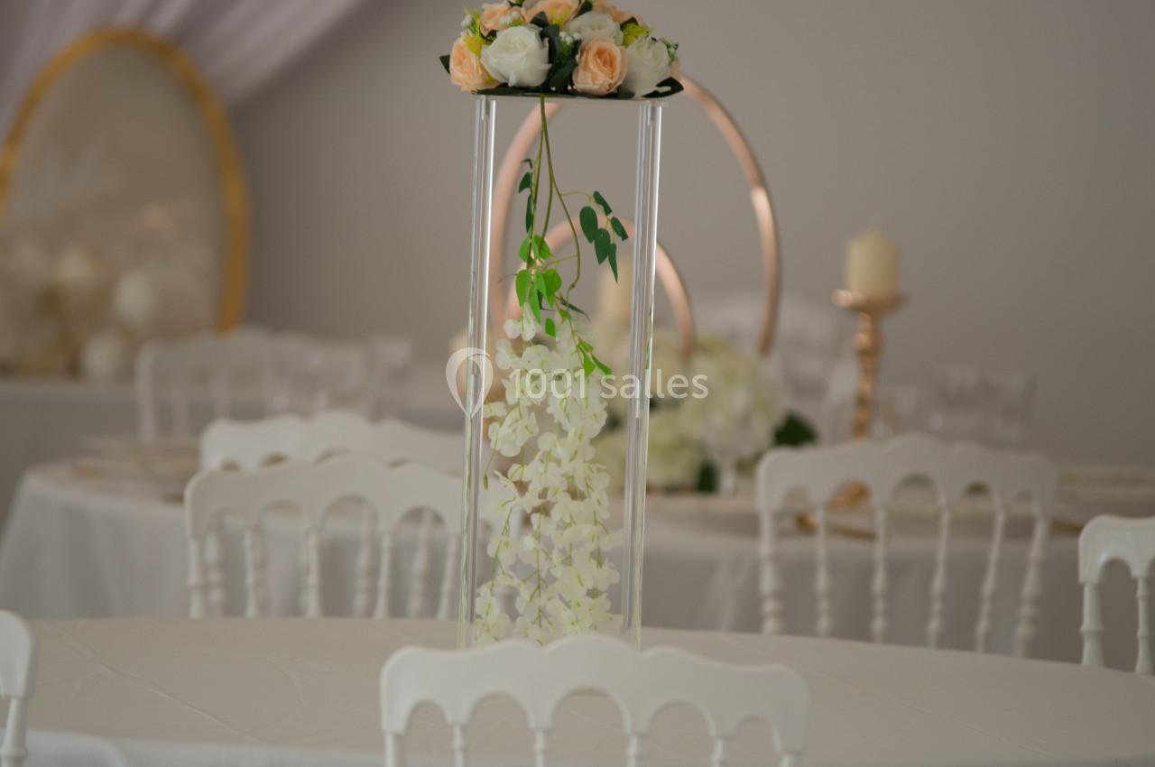 Centre de table floral avec roses et orchidées blanches, posé sur un support transparent dans une salle décorée.