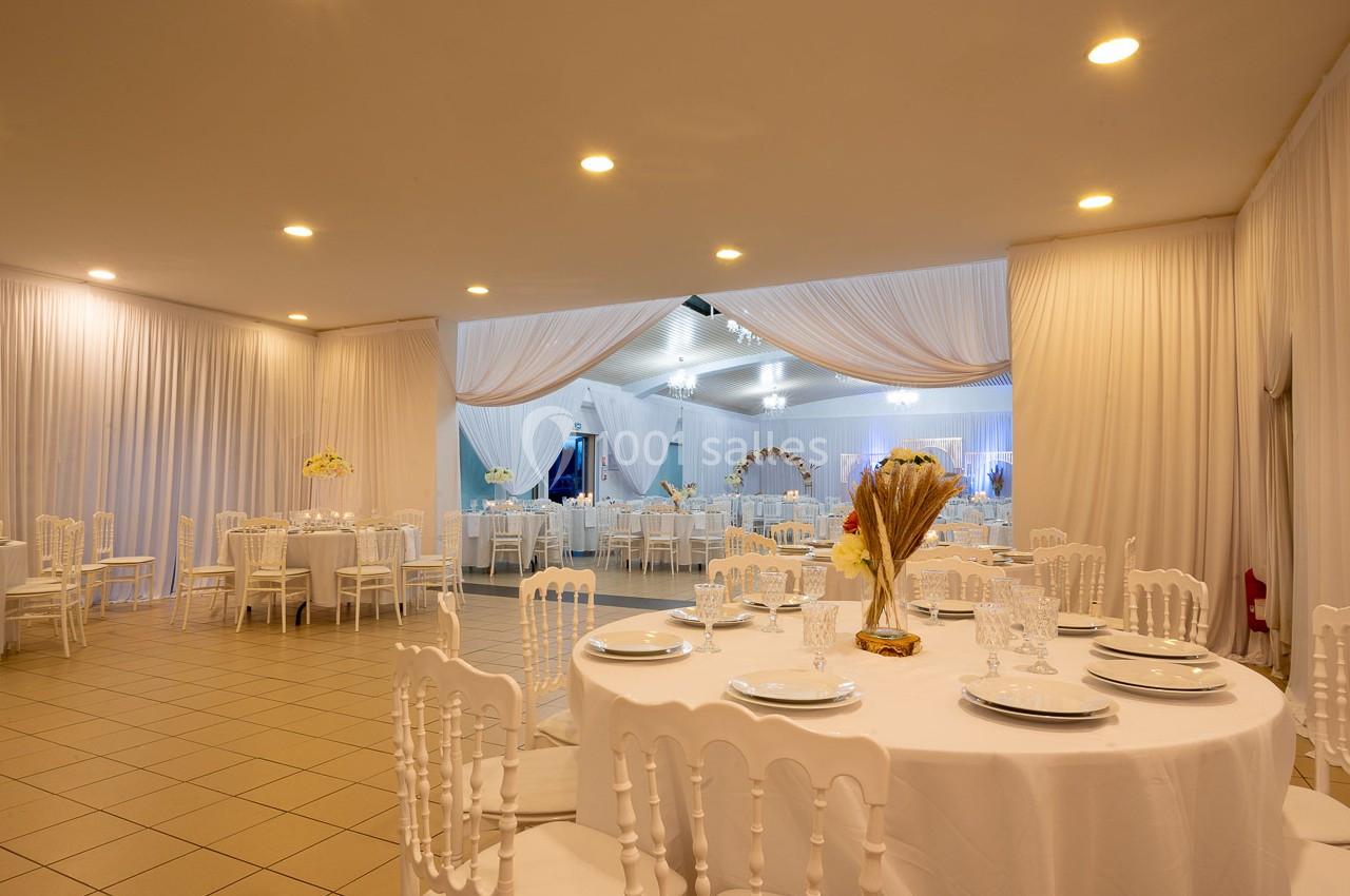 Salle de réception élégante avec tables rondes dressées, chaises blanches et rideaux en tissu clair.