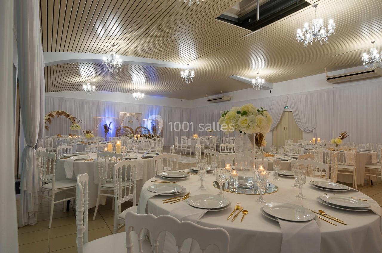 Salle de réception élégamment décorée avec des tables rondes, nappes blanches, chandeliers et éclairage chaleureux.