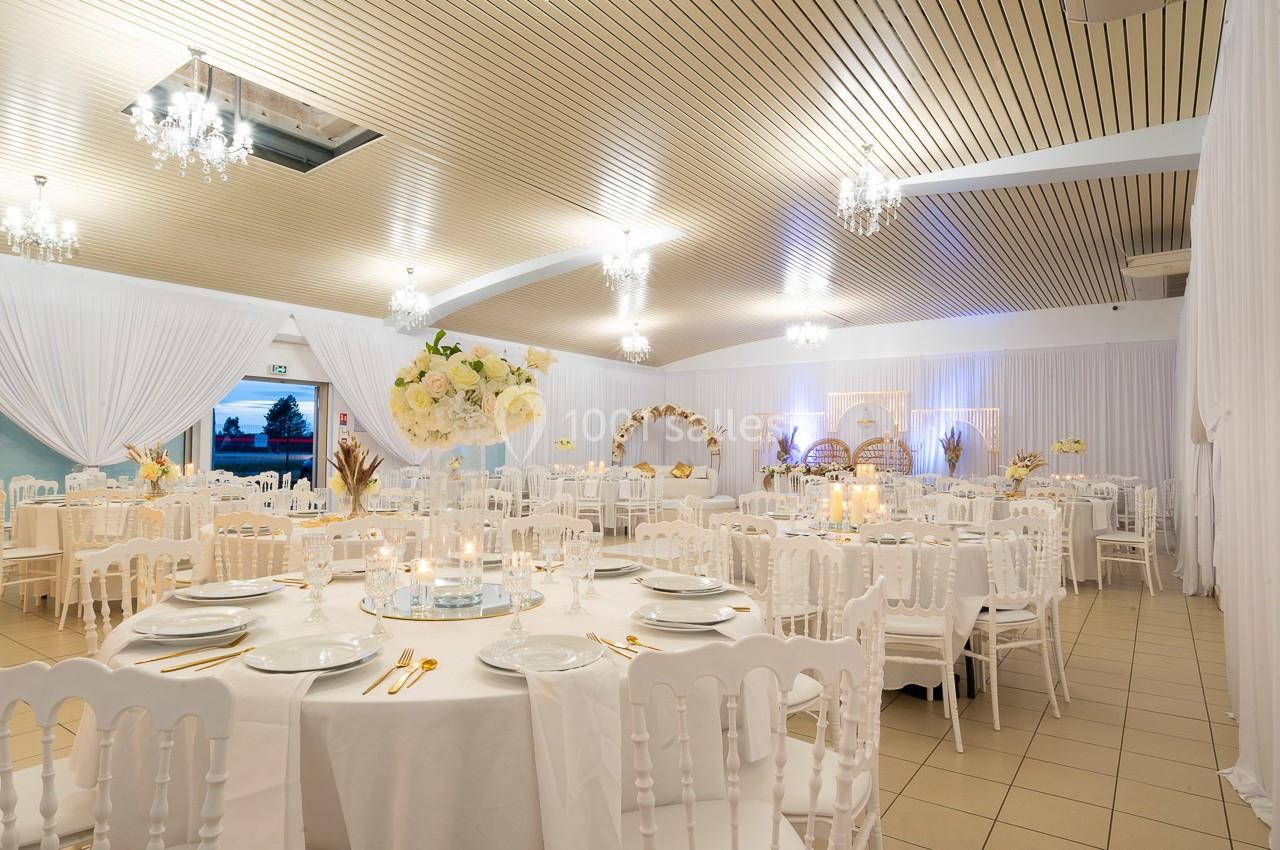 Salle de réception élégamment décorée avec des tables rondes, des chaises blanches et des arrangements floraux.