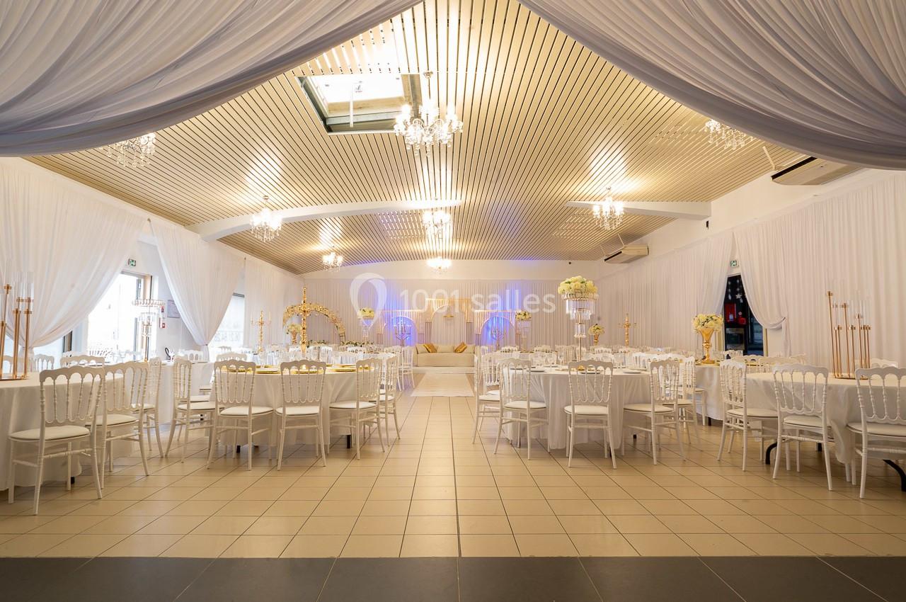 Salle de réception élégante avec tables rondes dressées, chaises blanches et décorations lumineuses sous un plafond en bois.