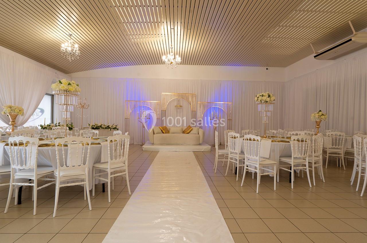 Salle de réception décorée avec des tables rondes, des chaises blanches et une estrade ornée de fleurs et de lumière.