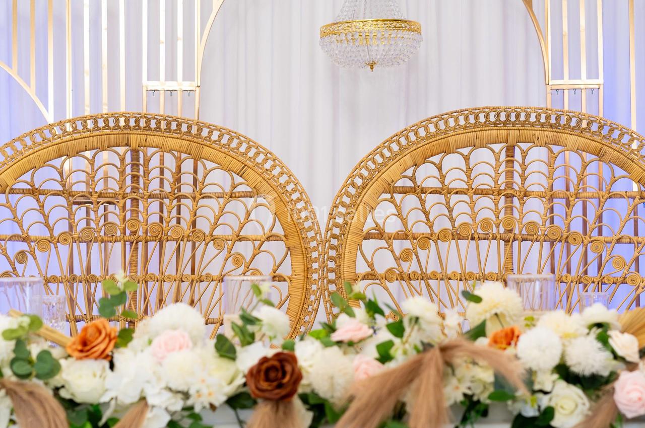 Deux fauteuils en rotin ornés de motifs complexes, devant une décoration florale et un rideau blanc avec un lustre suspendu.