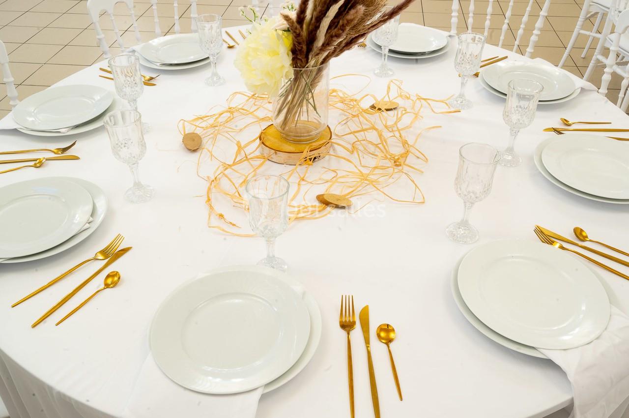 Table ronde dressée avec vaisselle blanche, couverts dorés, verres en cristal et centre de table décoratif avec fleurs…