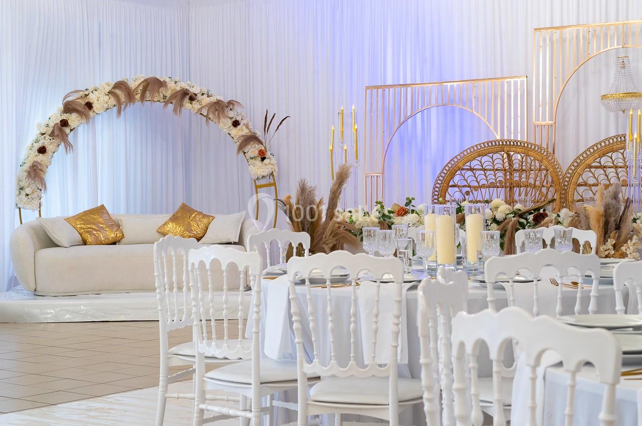 Salle décorée pour un événement avec arche florale, fauteuils dorés, chandeliers et table dressée avec chaises blanches.