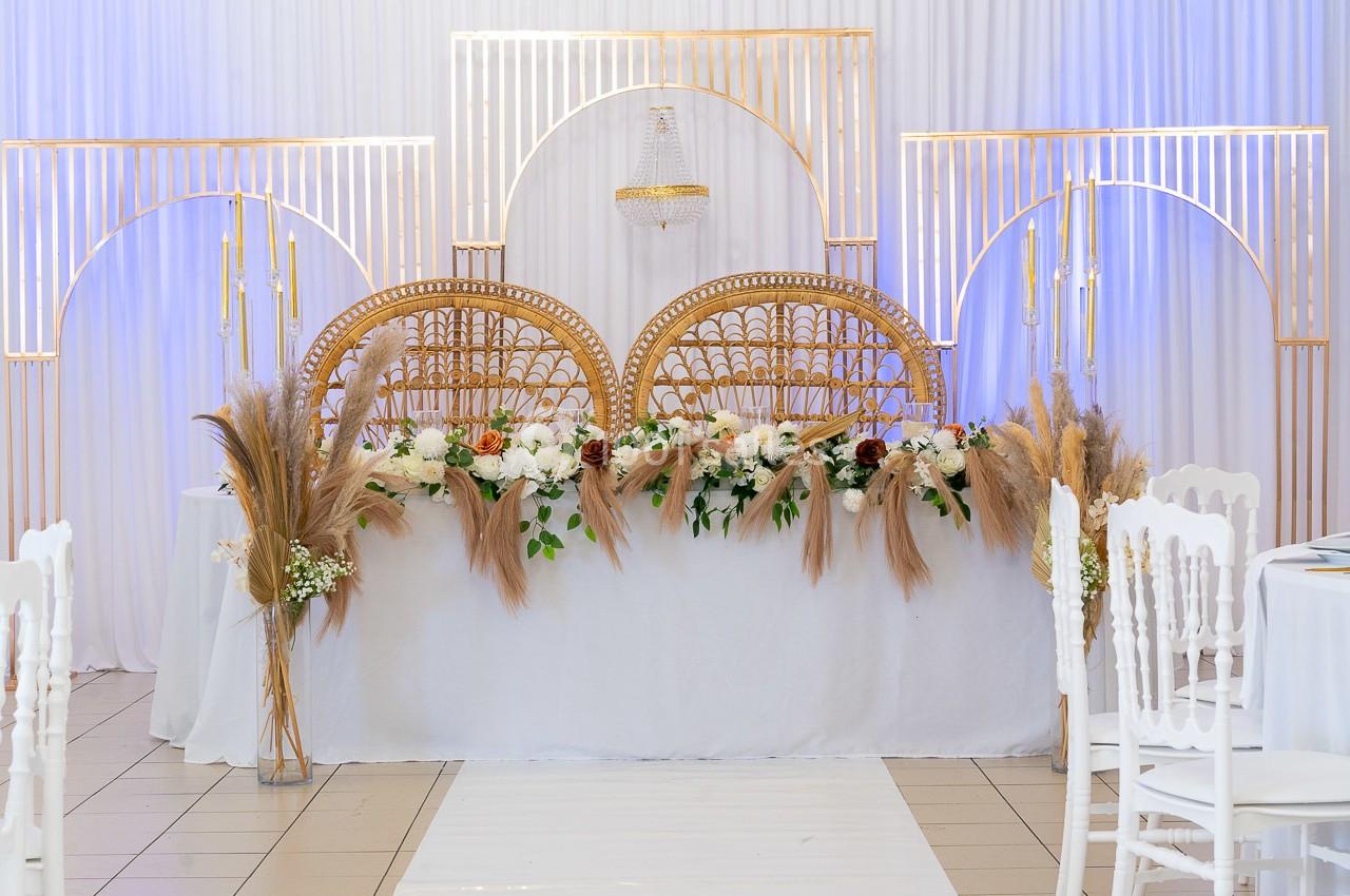Décoration de table de mariage avec chaises en rotin, fleurs blanches et pampas, devant un fond blanc et doré.