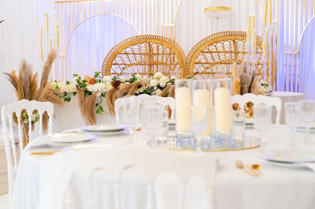 Table dressée avec vaisselle blanche au premier plan, décor floral et chaises en rotin doré en arrière-plan.