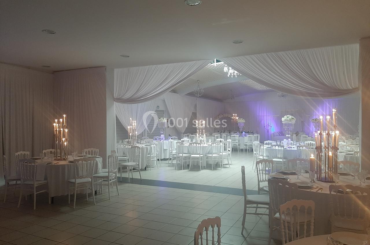 Salle de réception décorée avec des tables rondes, des chaises blanches, des chandeliers et des drapés blancs au plafond.
