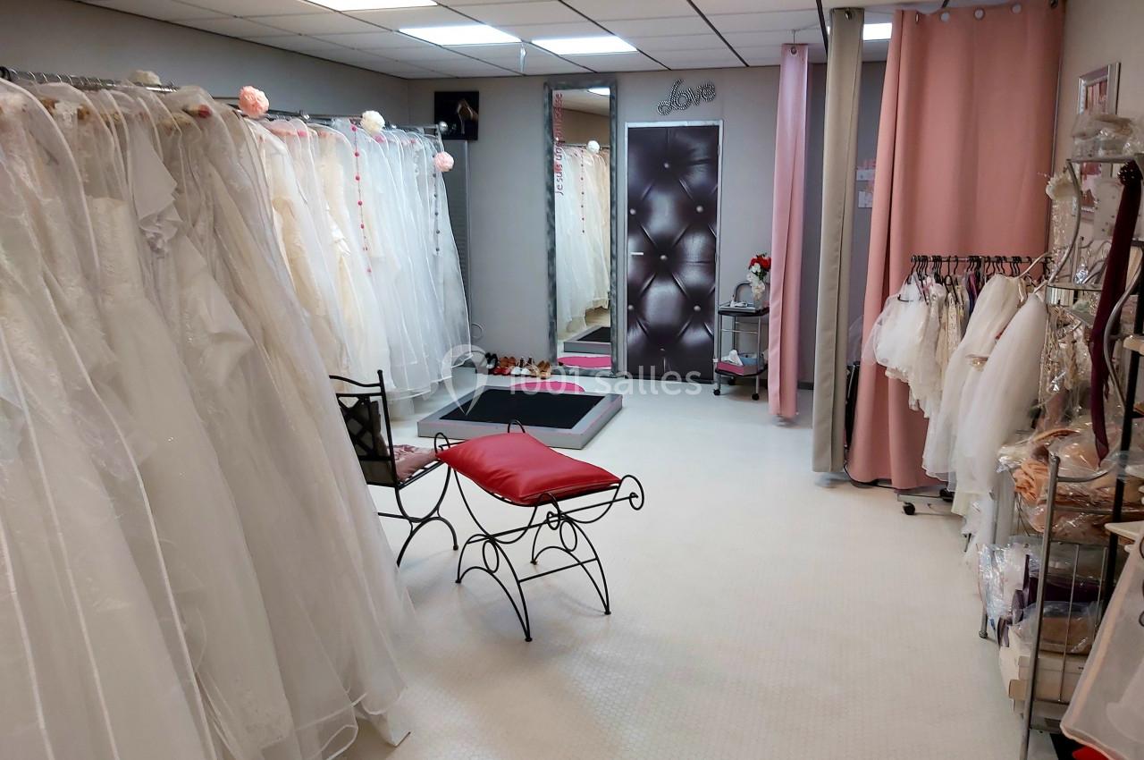 Salle d'essayage avec robes de mariée suspendues, miroir, fauteuil rouge et rideaux roses.