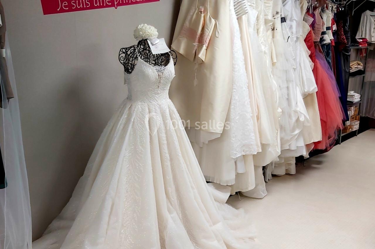 Robe de mariée blanche exposée dans une boutique, entourée d'autres tenues suspendues sur des portants.