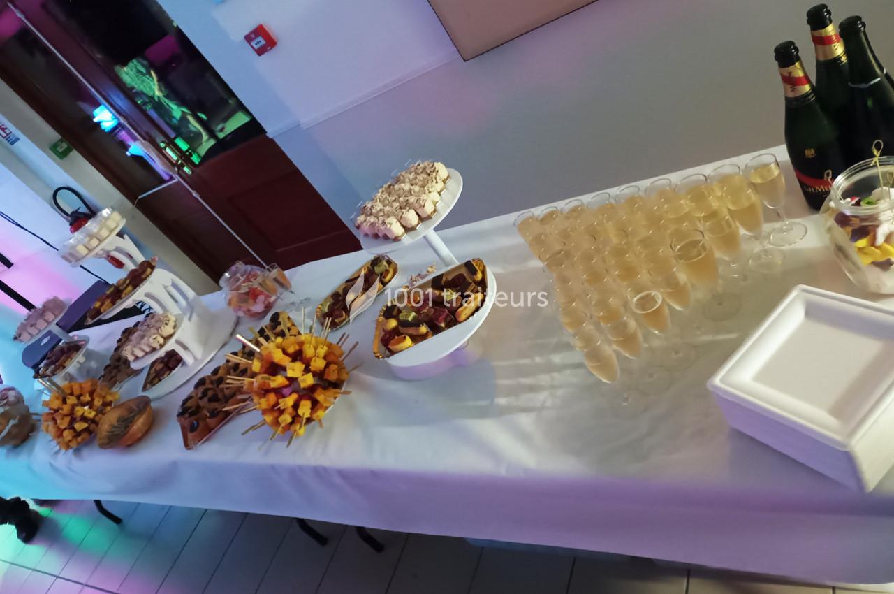 Buffet avec verrines, brochettes de fruits, pâtisseries et bouteilles de champagne sur une table blanche.