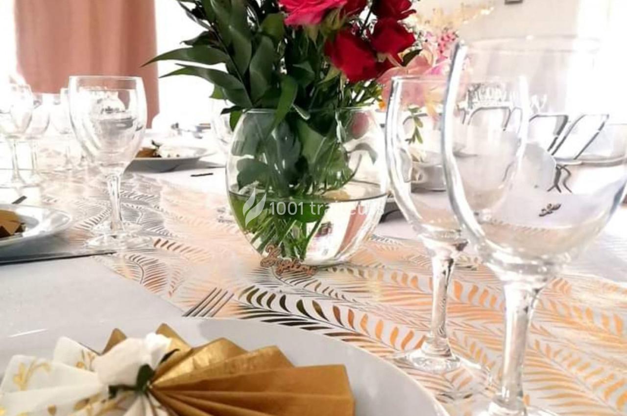 Table dressée avec des verres, des serviettes dorées pliées et un vase contenant un bouquet de fleurs rouges et roses.