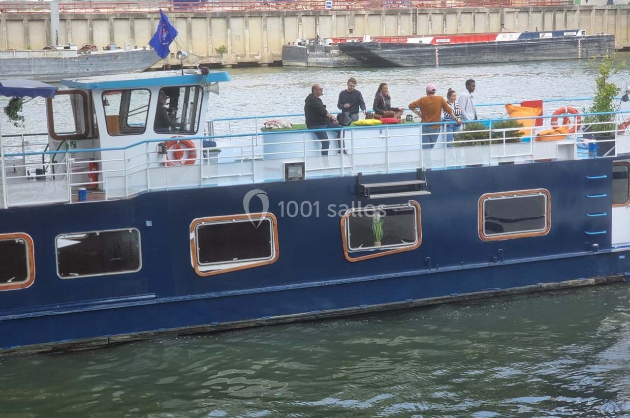 Bateau bleu amarré sur un canal, avec des personnes discutant sur le pont supérieur.