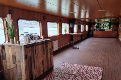 Intérieur d'une péniche aménagée avec un parquet en bois, un bar en bois, des plantes et des sièges décorés de coussins.